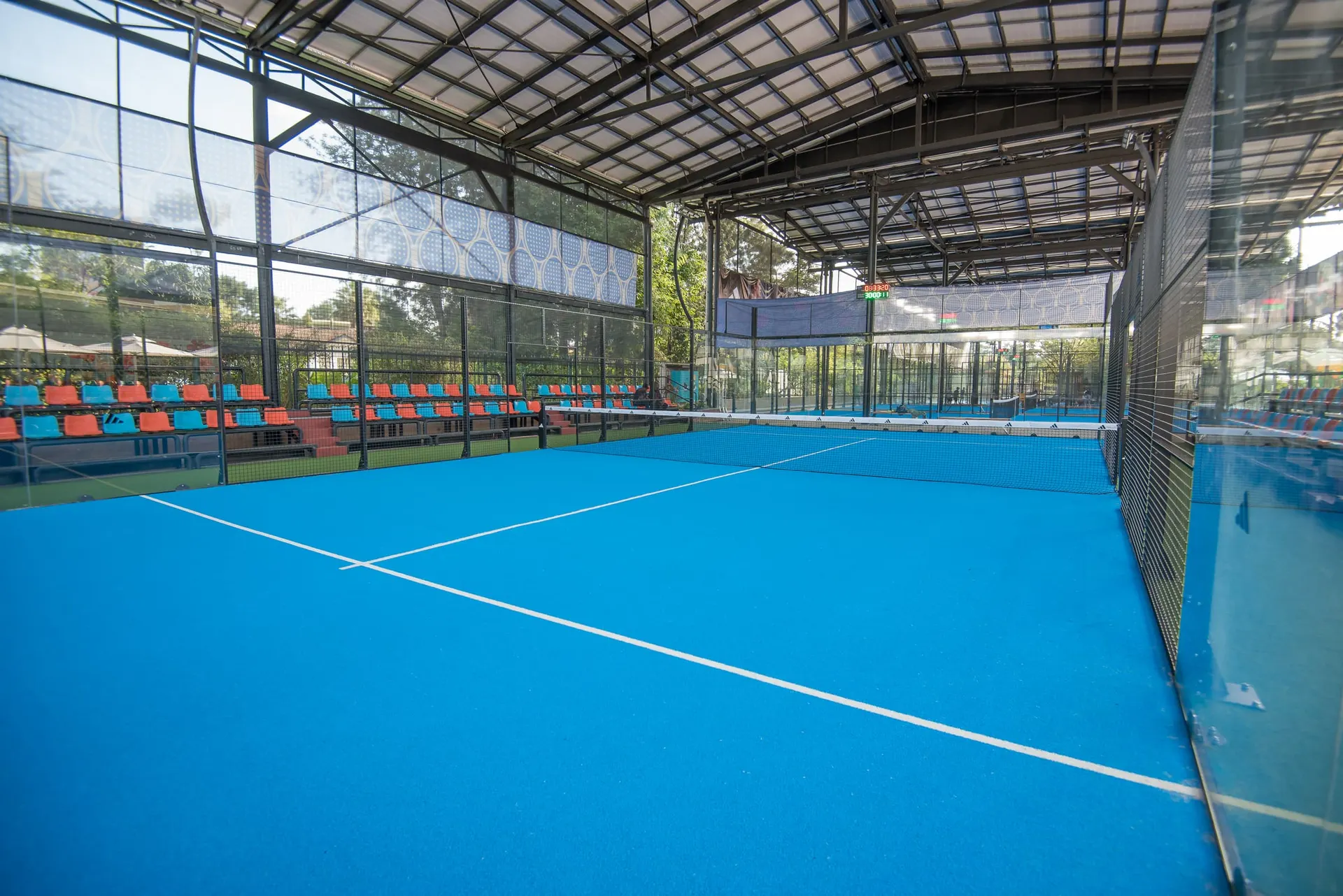 A padel court beneath the steel-framed roof, spectator seating alongside.