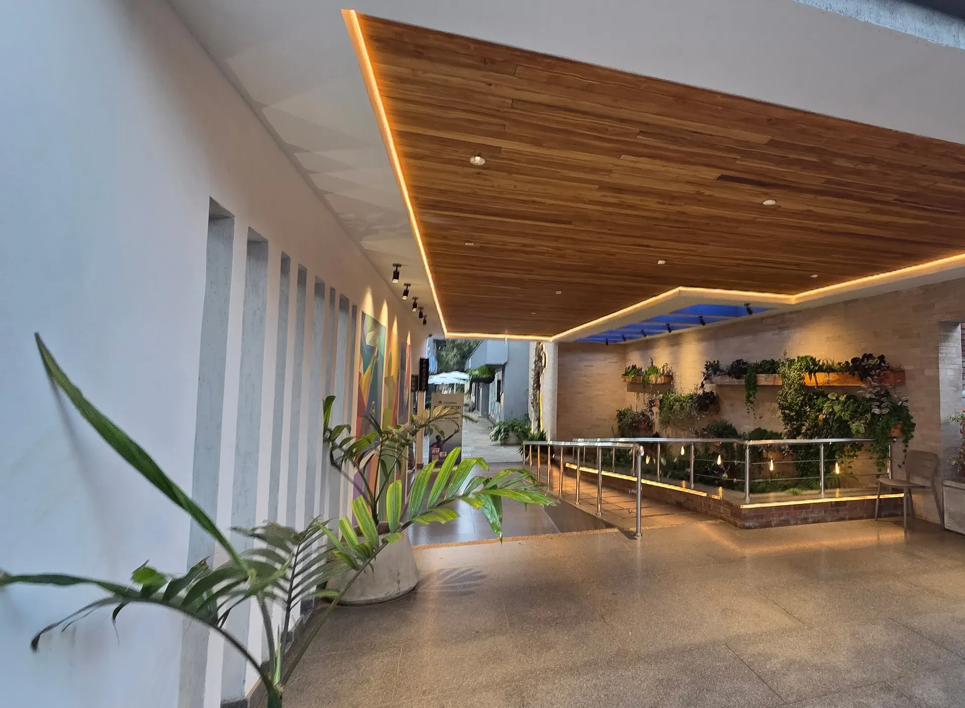 The arrival colonnade at Networks Padel Village: a faceted timber soffit lit by a continuous cove light, a planted wall and brick planter beyond, terrazzo underfoot.